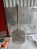 Full view of the pizza peel standing against a metal surface showing wooden handle and aluminum blade.