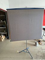 Front view of the vintage Da-Lite projection screen fully extended on tripod stand, showing the gray screen surface and blue edges.