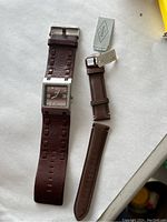 Roots watch with attached wide chocolate brown leather band and separate Fossil leather watch band with tags on a white background.