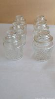 Six clear cut glass jars with detailed pattern and embossed rose lids arranged in two rows of three on a beige background.