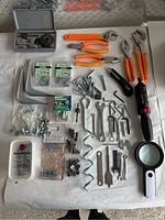Full view of all tools and hardware arranged neatly on a white surface, includes pliers, wrenches, cordless screwdriver, magnifier, and assorted screws/anchors.