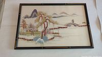 Frontal view of the framed embroidered artwork showing the Asian mountain village scene with houses, trees, bridge, and mountains.