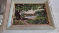 Framed oil painting depicting a landscape with a large tree, river, and bridge under daylight.
