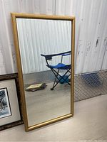 Full view of wood framed rectangular mirror standing on floor against wall, showcasing medium tone natural wood frame and clear reflective mirror surface.