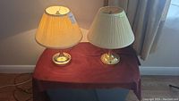 Two table lamps on a small table covered by a burgundy cloth, one lamp lit and the other not. Both with brass round bases and pleated cream fabric shades.