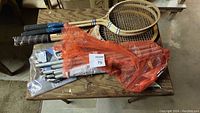 Photo of 3 vintage wooden badminton rackets stacked on a table with metal net poles and orange mesh net bag.