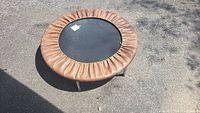 Brown small trampoline taken outdoors on a pavement, showing top view with brown protective padding around black jumping surface.