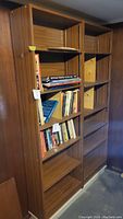 Double connected wooden bookcase unit in basement setting, one side with books, one side with empty shelves and compartments.