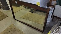 Large wooden framed mirror leaned against wall on concrete floor in storage area. Shows dark wood frame and some wear on frame.