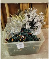 Multiple strings of diverse holiday Christmas lights with colored bulbs stored in a plastic bin along with fake foliage including white flowers and golden leaves