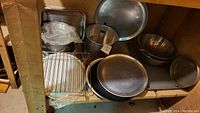 Shelf showing various metal bakeware including round perforated pizza pans, tube cake pan, mixing bowls, and cooling racks.