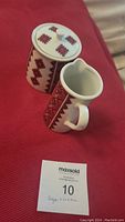 Angle view of sugar container with lid and creamer placed on red fabric. Shows geometric Ukrainian pattern in red and black on cream-colored ceramic.
