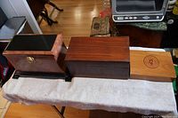 Photo showing all three wooden boxes side by side on a white towel-covered surface. Visible features include the light wood cigar box with Rembrandt logo, dark brown rectangular mahogany box, and the faux wood box with gold lady face detailing.