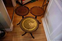 Three small wooden tables with tripod legs arranged on floor, two with round tops featuring gold edging and one with scalloped wood top.