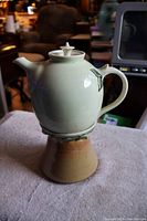 Full view of the signed ceramic tea pot with matching warmer stand beneath it, made of glazed pottery with floral details.
