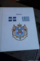 Front cover of the Greece stamp album showing two Greek flags and a coat of arms with two figures on either side.