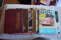 Top view showing the brown telephone/address book on top, Popular Science magazine with a car on its cover, and several other documents or books underneath on a light tablecloth.