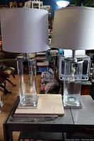 Pair of tall clear cylindrical glass lamps with off-white drum shades placed on a wooden surface in a room with furniture in the background.