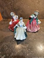 Three Royal Doulton porcelain figurines of women in elegant dresses and bonnets on a dark surface.