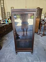 Front view of tall antique English style bookcase with glass door closed, showing dark wood frame and small wheels at bottom.