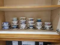 Full view of 16 cups and saucers displayed on two shelves inside a wood cabinet