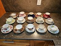 Overall view of the 15 assorted bone china cups and saucers arranged on a countertop showing the variety of patterns and colors.