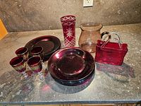 Overview photo showing 8 cranberry glass plates stacked, 4 sherry glasses with gold trim, cranberry etched hurricane vase, clear glass pitcher, and basket vase with handle.
