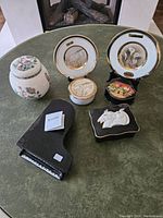 Seven decorative trinket boxes and plates arranged on table showing all items including plates with gold rims, floral jar, village painted black box, and piano shaped box