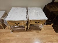 Pair of end tables side-by-side with visible white quartz tops and beige/carved wooden bases wrapped partially in plastic.
