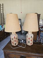 Pair of mid century table lamps shown side by side on wooden furniture, beige fabric lampshades, ceramic textured bases with red and black colored insets.