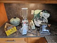Photo of multiple retro kitchen appliances arranged on a kitchen countertop including a green EUREKA electric wok, blender, two hand mixers, toaster, and box of vintage utensils