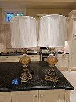 Pair of French style table lamps with white drum shades on black countertop in kitchen setting.