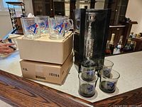 Photo showing eight Toronto Blue Jays glass cups on cardboard boxes, four tumblers with Blue Jays logo in front, and silver Maple Leaf Stanley Cup bank in plastic display case behind.