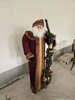 Full view of tall Santa figure, showing full coat, fur trim, and garland with ornaments and bows.