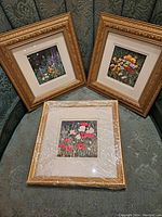 Trio of three framed floral paintings displayed on green patterned upholstery, one wrapped in plastic.