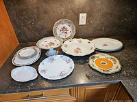 Photo showing the assortment of fine bone china plates, pedestal cake stand, and serving bowls with various floral patterns and gold accents arranged on a dark countertop.