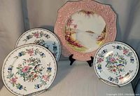 Four plates arranged with three Aynsley Pembroke plates in front, showing colorful floral and bird designs, and one large Royal Winton pink embossed plate with water scene behind them.