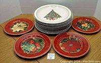 Set of 12 Christmas decorative plates displayed on a wooden surface, 7 white plates with Christmas tree design stacked and 5 red border plates spread out