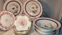Stacked Royal Dux and Paragon Minuet plates with floral and bird designs. Front showing detailed floral patterns and bird motifs with burgundy and gold trim.