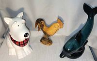 Three miscellaneous figures: white ceramic dog treat jar shaped as a dog wearing a plaid scarf, yellow wooden carved rooster, and blue-green glazed dolphin.