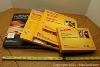 Four items arranged on a wooden surface with measuring tape showing approximately 13 inches width. Items include three unopened photo paper packages and one hardcover book titled 'Step-by-Step Picture Framing' by Ken Kandler.