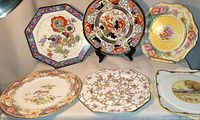 Six vintage decorative plates arranged on display showing various floral and scenic patterns with different shapes including round, octagonal, and square.
