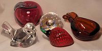 Five glass items including cranberry glass apple, clear glass ball paperweight with colorful internal details, clear glass bird, red strawberry-shaped glass, and dark amber Murano pear-shaped ashtray.