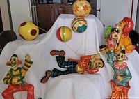 Five-piece lot of paper mâché clowns and lion, all colorful, placed on white cloth draped surface. Clowns are holding balls on sticks and strings, lion with orange mane and green parrot figure on its back.