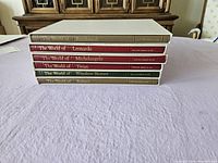 Stack of six art books on a purple tablecloth, titles from classic art masters visible on book spines: Rembrandt, Leonardo, Michelangelo, Titian, Winslow Homer, Rubens.