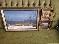 Photo showing all three framed artworks on a green upholstered background: one large winter landscape and two smaller miniature paintings