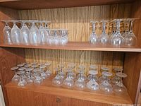 Shelf displaying multiple rows of clear stemware glasses including water, liquor, wine, coupe, and champagne glasses all placed upside down.