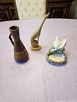 Three vintage ceramic pieces displayed on a light purple tablecloth including the pair of geese ashtrays, brown vase, and tan bird figurine vase.