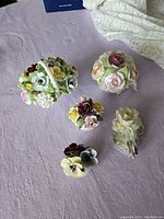 Five porcelain flower figurines displayed on a lavender cloth including a floral basket and multiple flower clusters.
