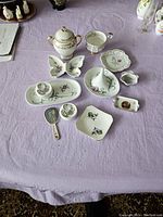 Full view of all 11 porcelain pieces arranged on a purple cloth surface including miniature mirror, lidded sugar bowl, various decorative dishes and trays